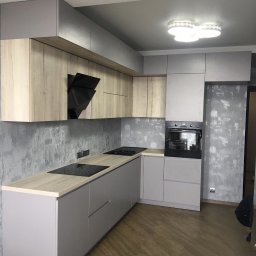 Max bud - Szara zabudowa kuchenna z drewnianymi akcentami, czarny okap i płyta indukcyjna na tle ściany z efektem betonu.