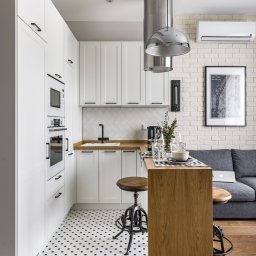 Max bud - Nowoczesna, jasna kuchnia z białymi szafkami, drewnianym blatem, chromowanym okapem, wzorzystą podłogą i ceglaną ścianą, widok na salon z szarą sofą.