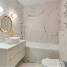 Nowoczesna łazienka z wanną obłożoną marmurowymi płytkami, umywalką nablatową z baterią w kolorze złota, ośmiokątnym lustrem w złotej ramie i grzejnikiem drabinkowym.