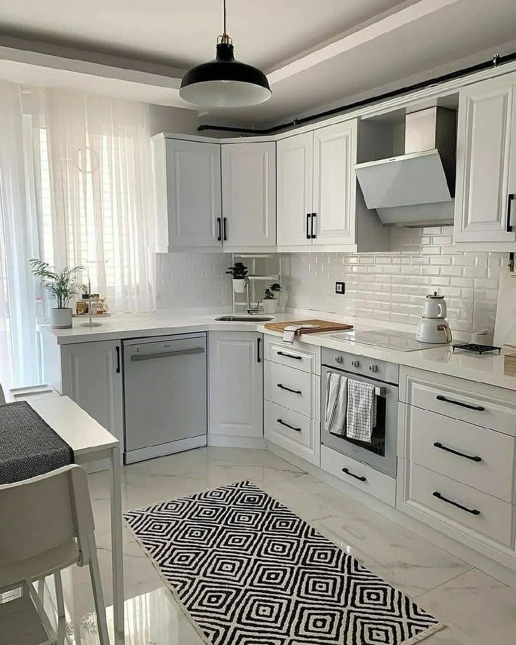 Jasna kuchnia w stylu skandynawskim z białymi szafkami i czarnymi uchwytami, białe płytki na ścianie, marmurowy blat, czarny dywan z geometrycznym wzorem, okno z firanką i czarna lampa sufitowa.