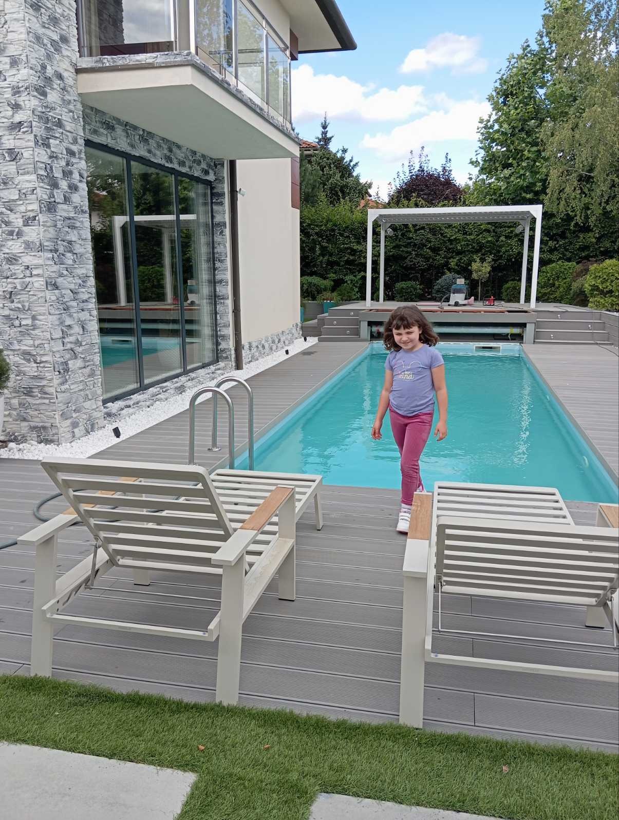Dziewczynka idzie brzegiem prostokątnego basenu z szarą drewnianą obudową, obok dwa białe leżaki, w tle nowoczesny dom z kamienną elewacją i dużymi oknami.