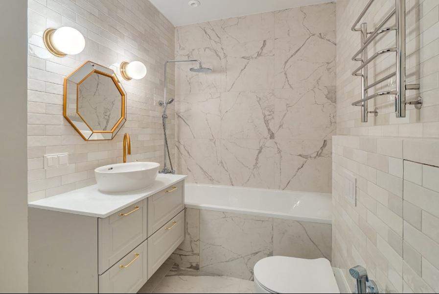 Nowoczesna łazienka z wanną obłożoną marmurowymi płytkami, umywalką nablatową z baterią w kolorze złota, ośmiokątnym lustrem w złotej ramie i grzejnikiem drabinkowym.