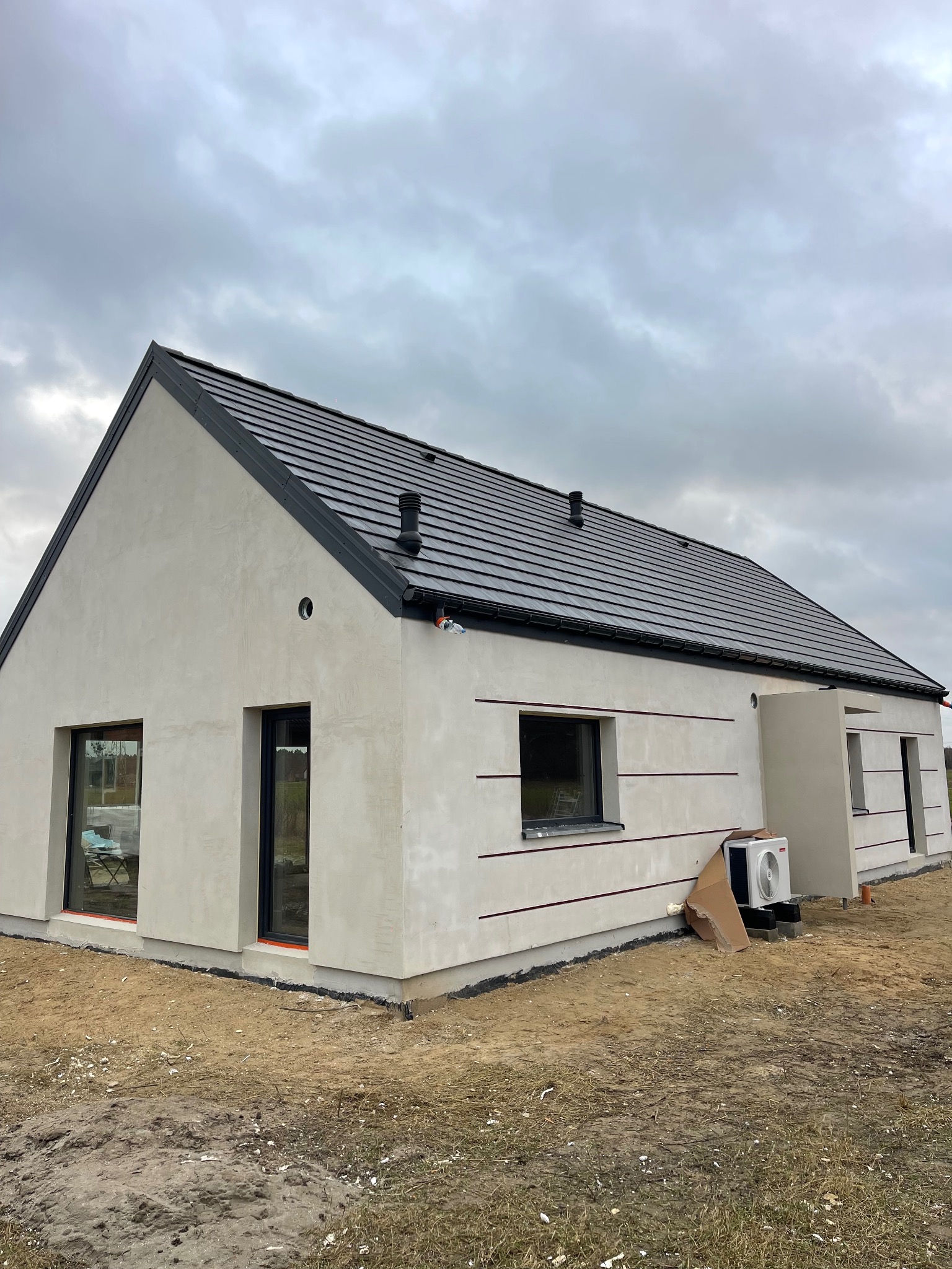 Surowa elewacja jednorodzinnego domu z czarną dachówką, widoczne poziome dekoracyjne frezy, klimatyzator zewnętrzny w kartonie, teren wokół domu częściowo zagospodarowany.