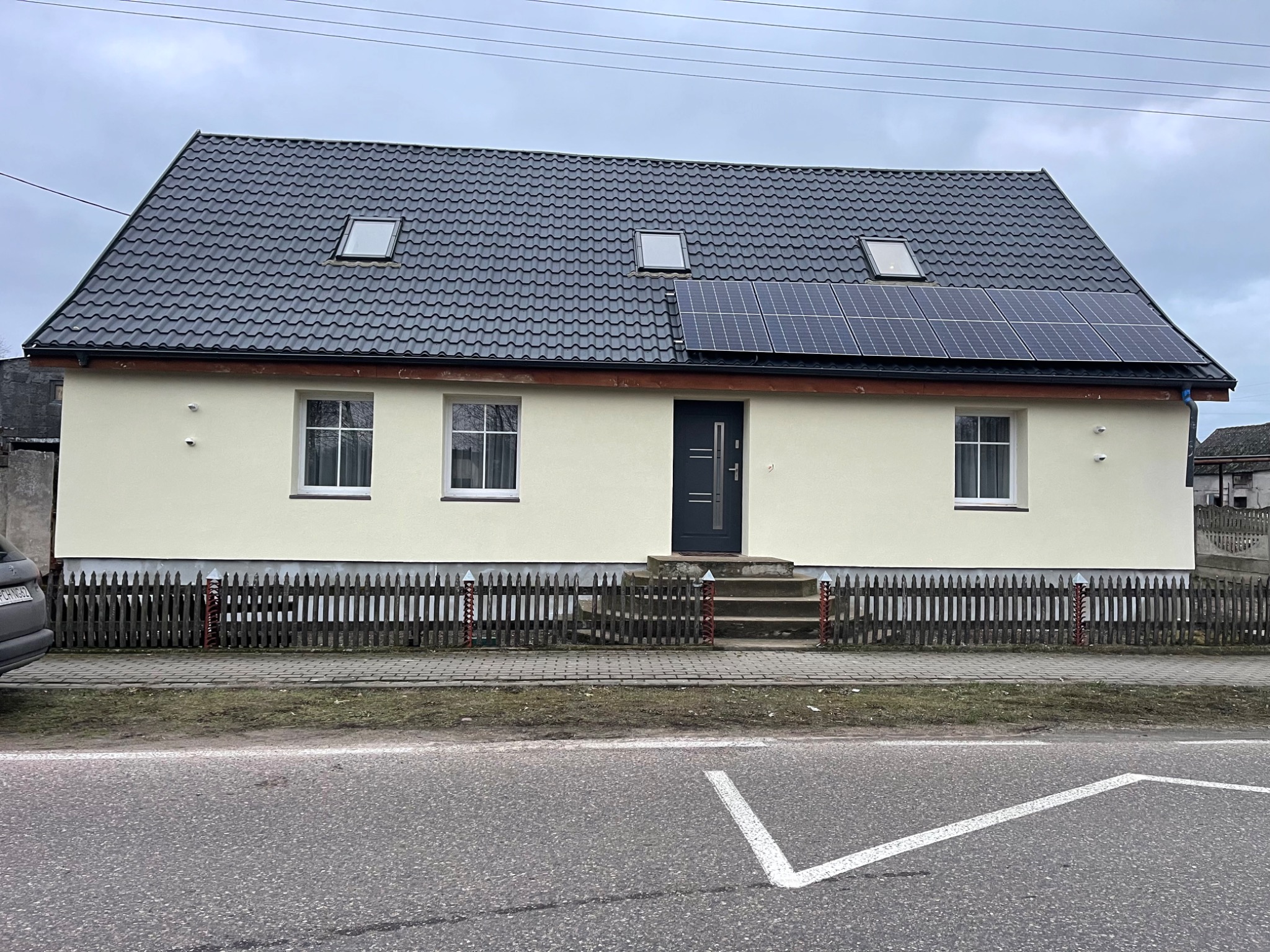 Dom jednorodzinny po remoncie elewacji, z panelami słonecznymi na dachu i nową stolarką okienną, widziany z poziomu ulicy w pochmurny dzień.