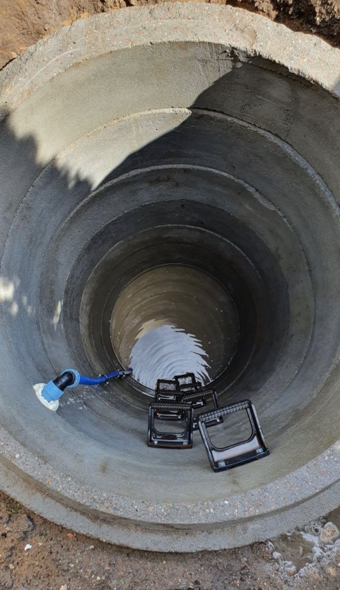Wnętrze betonowej studni kręgowej z wodą na dnie, widoczny niebieski wąż podłączony do pompy i czarne stopnie drabinkowe.