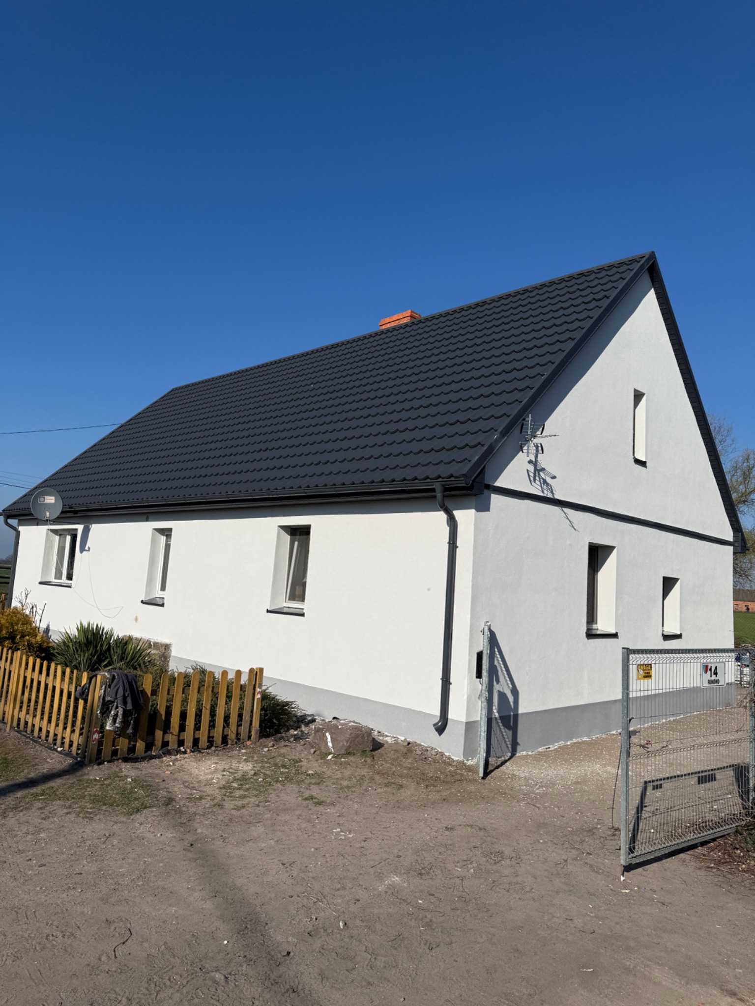 Biały dom z czarnym dachem po remoncie elewacji, widoczna antena satelitarna i brama wjazdowa z numerem posesji
