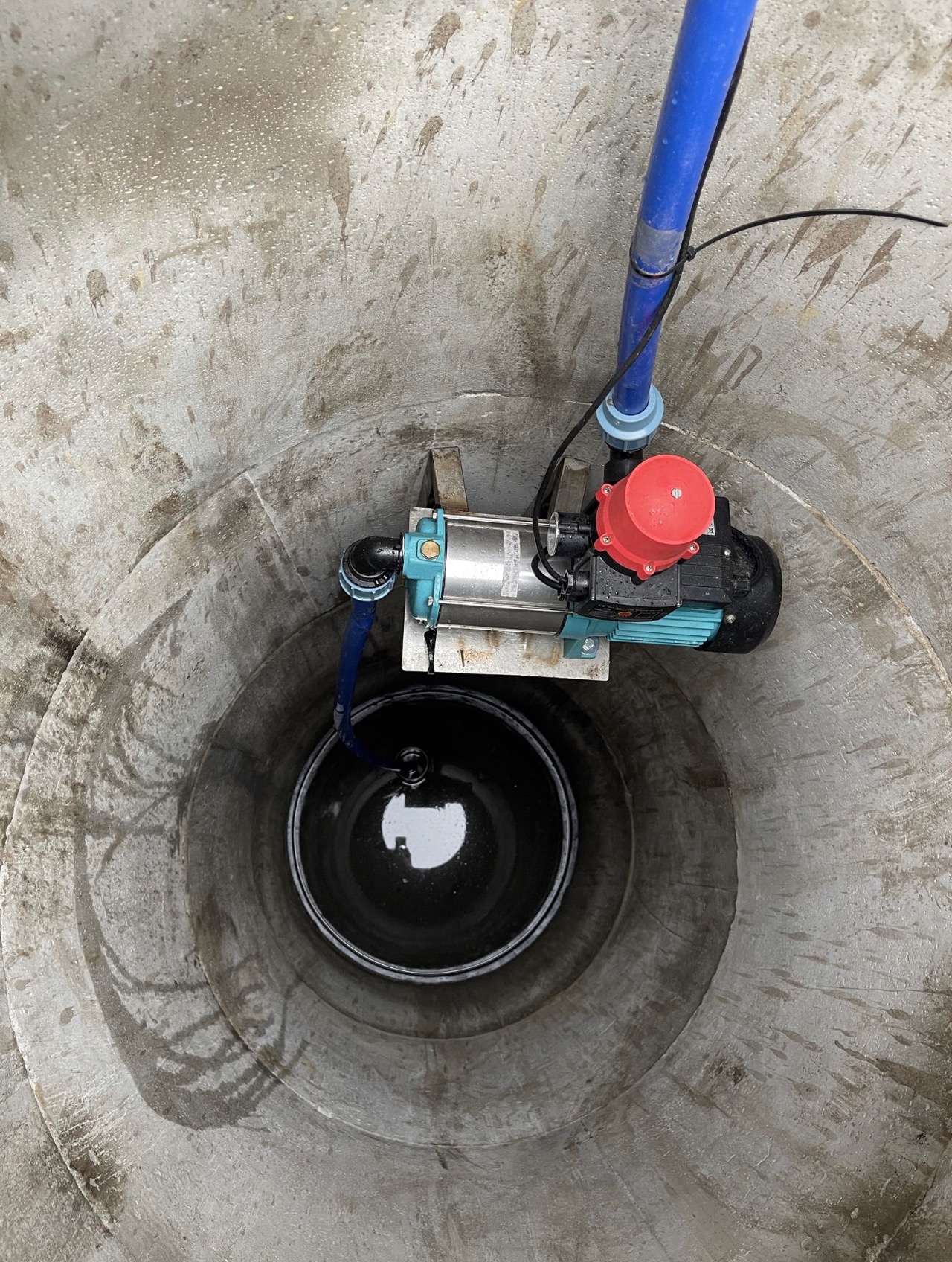 Wnętrze betonowej studni kręgowej z zainstalowaną pompą hydroforową, widoczne ślady wilgoci na ścianach i odbicie światła w lustrze wody.