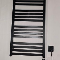 Pawel Zarjewski - Czarny, elektryczny grzejnik łazienkowy drabinkowy, zamontowany na białej ścianie, z widocznym gniazdkiem i spiralnym kablem zasilającym.