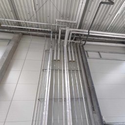 Pawel Zarjewski - Równoległe rury instalacyjne, niektóre zaizolowane, prowadzone wzdłuż ściany w hali, z widocznymi elementami podwieszenia pod sufitem. Metalowa konstrukcja.