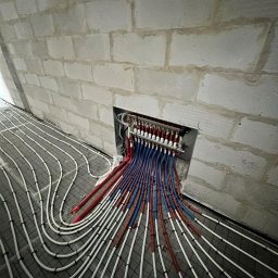 Pawel Zarjewski - Rozdzielacz ogrzewania podłogowego z rurami rozprowadzonymi na siatce w surowym wnętrzu. Rury białe, niebieskie i czerwone. Ściana z białych bloczków.