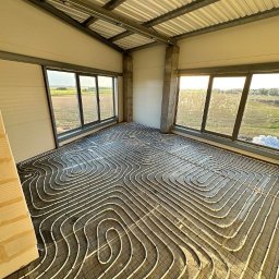 Pawel Zarjewski - Białe rury ogrzewania podłogowego ułożone na siatce w jasnym pomieszczeniu z oknami i widokiem na pole. Metalowa konstrukcja dachu, surowy stan wykończenia.