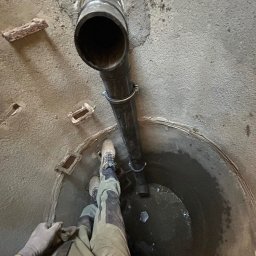 Pawel Zarjewski - Wnętrze studni kanalizacyjnej z zamontowaną pionową rurą odpływową, zabezpieczoną obejmami. Widoczne dno studni z wodą i fragment sylwetki pracownika w odzieży roboczej.