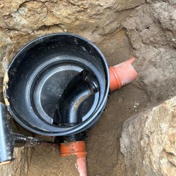 Pawel Zarjewski - Czarna studzienka kanalizacyjna z rurami w wykopie, widok z góry. Element instalacji kanalizacyjnej w ziemi, pomarańczowe rury. But na dole kadru.