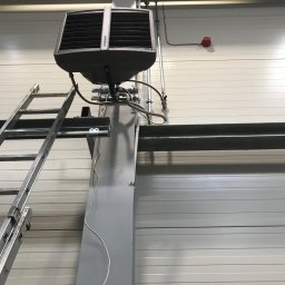 Pawel Zarjewski - Szary nagrzewnica powietrza Sonniger zamontowana na metalowej kolumnie, drabina oparta o kolumnę, instalacja elektryczna i przeciwpożarowa na ścianie z paneli.