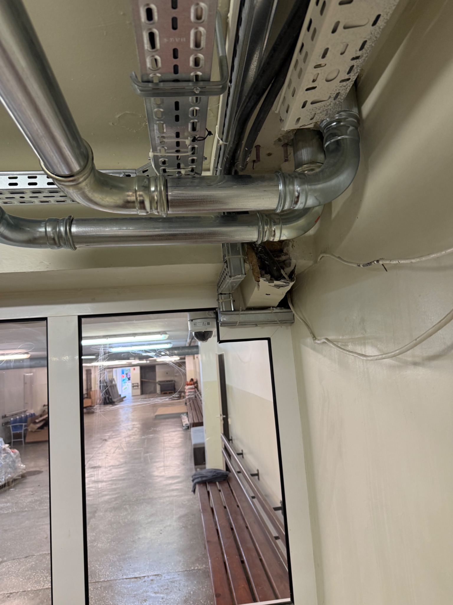 Instalacja rur i kabli pod sufitem, widoczne metalowe rury, korytka kablowe i uszkodzona obudowa. W tle widoczne pomieszczenie przez szklane drzwi.