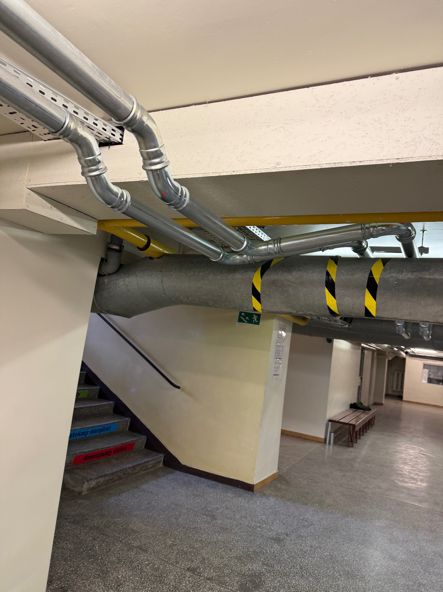Szare i metalowe rury instalacyjne pod sufitem w korytarzu. Widoczne elementy mocowań, rury z żółto-czarnymi pasami, schody z napisami w tle.