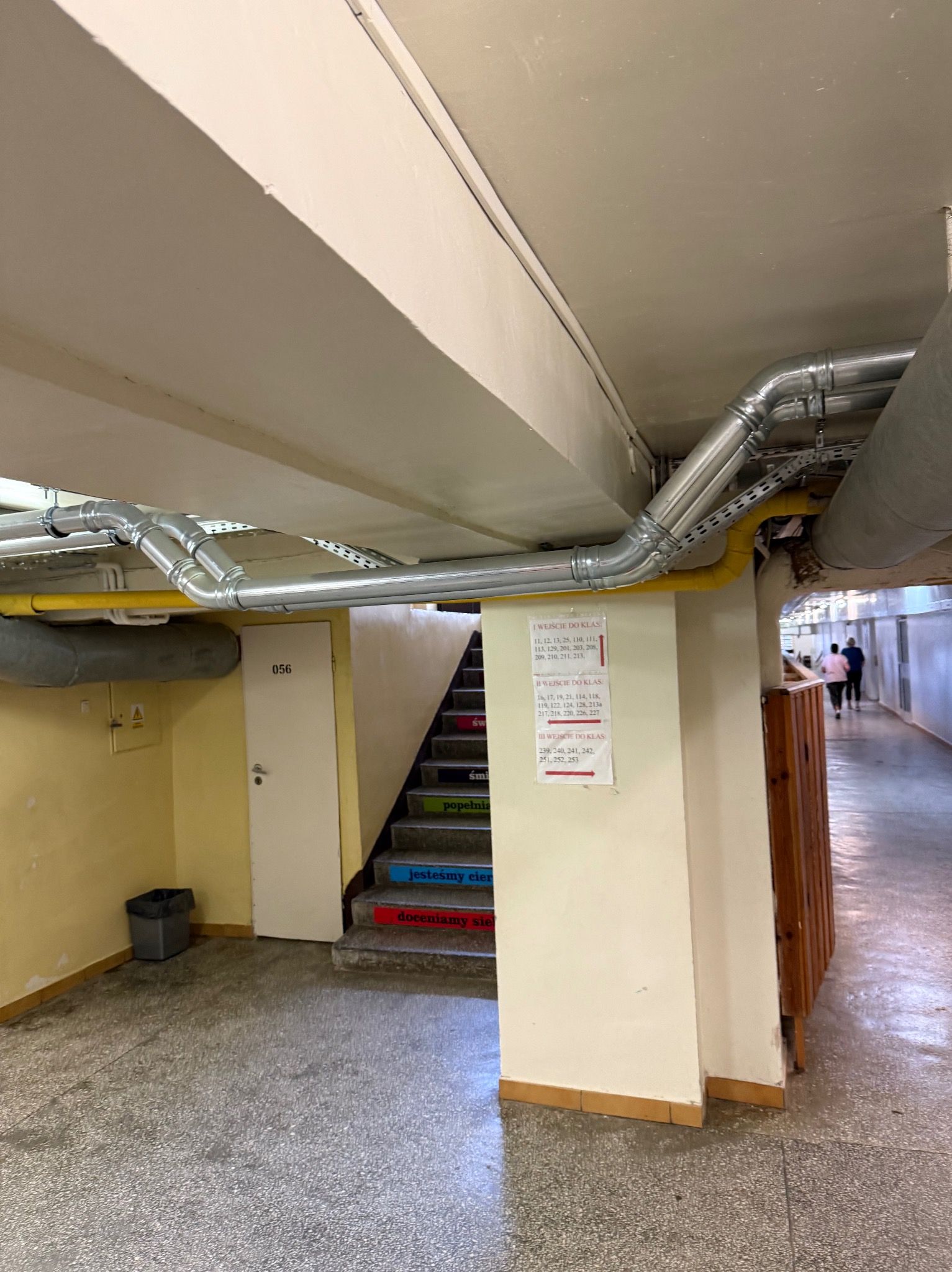 Instalacja rur kanalizacyjnych pod sufitem w obiekcie w Olsztynie. Rury stalowe, kolanka, mocowania. Widoczne schody i tablica z numerami sal.