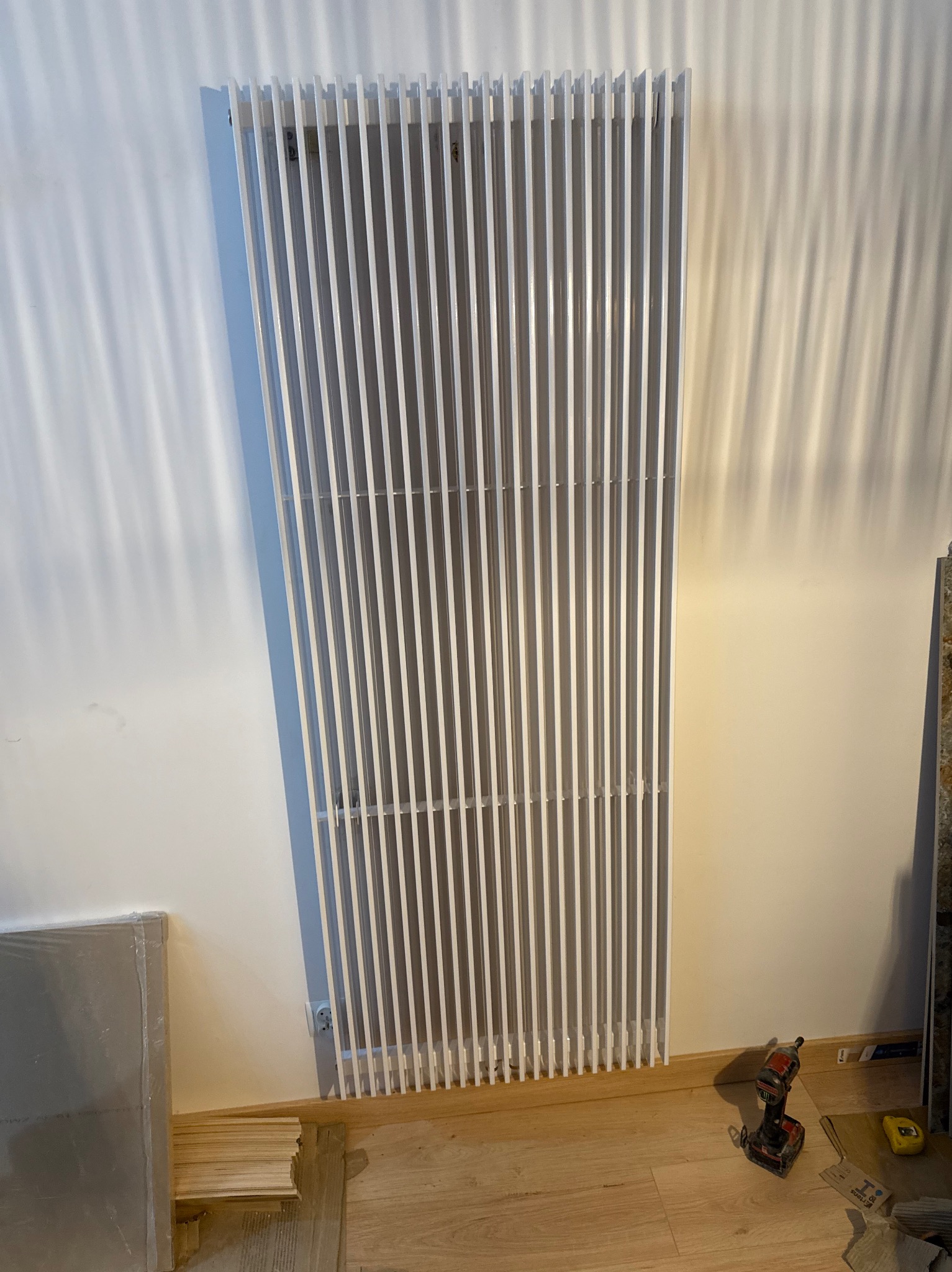 Biały, dekoracyjny grzejnik panelowy pionowy, zamontowany na ścianie. Widoczne narzędzia i materiały montażowe. Nowoczesny design, minimalistyczne wnętrze w trakcie wykończenia.
