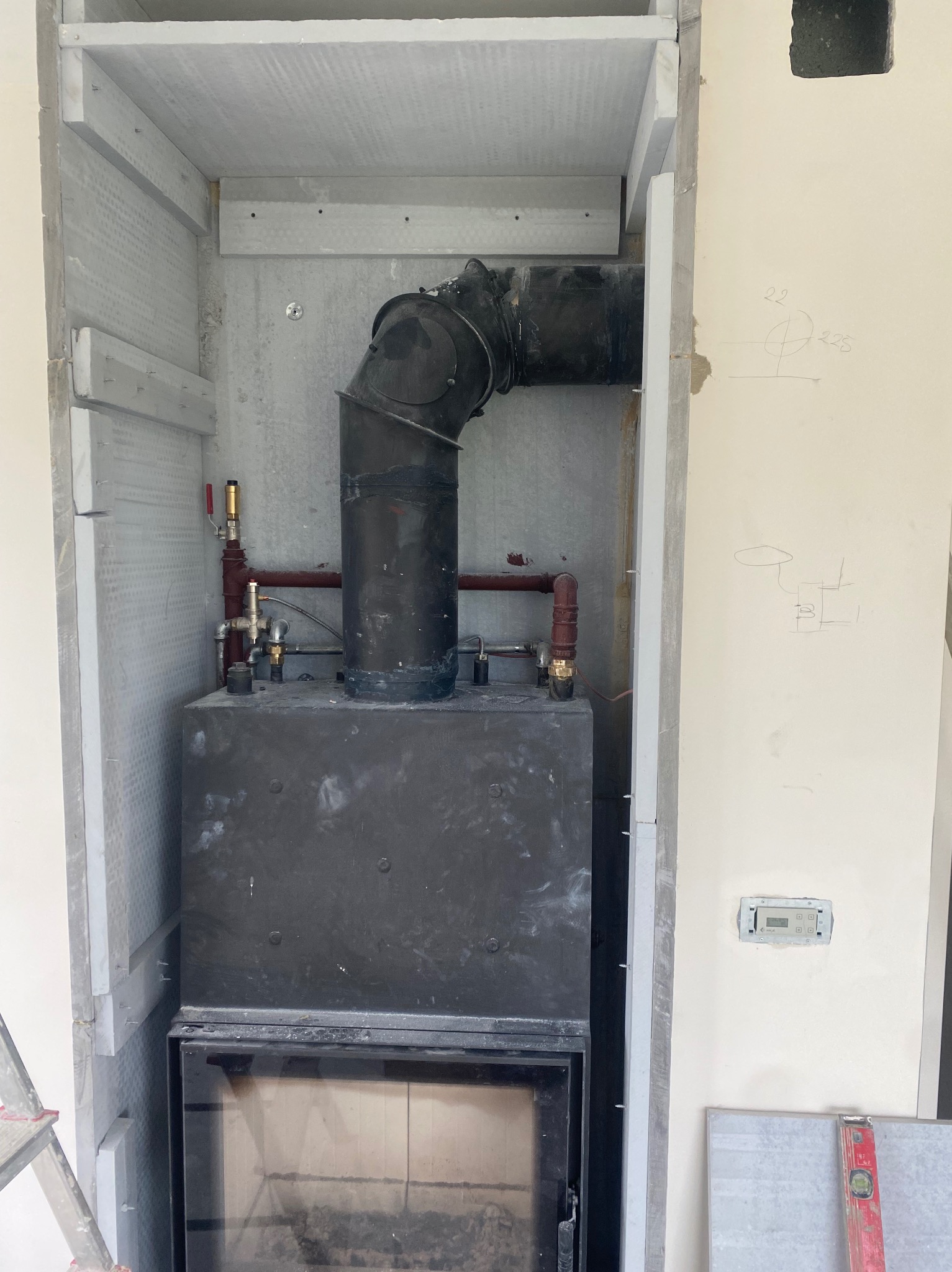 Czarny piec z rurą w zabudowie z płyt. Widoczne elementy instalacji i fragment drabiny. Na ścianie szkic instalacji.