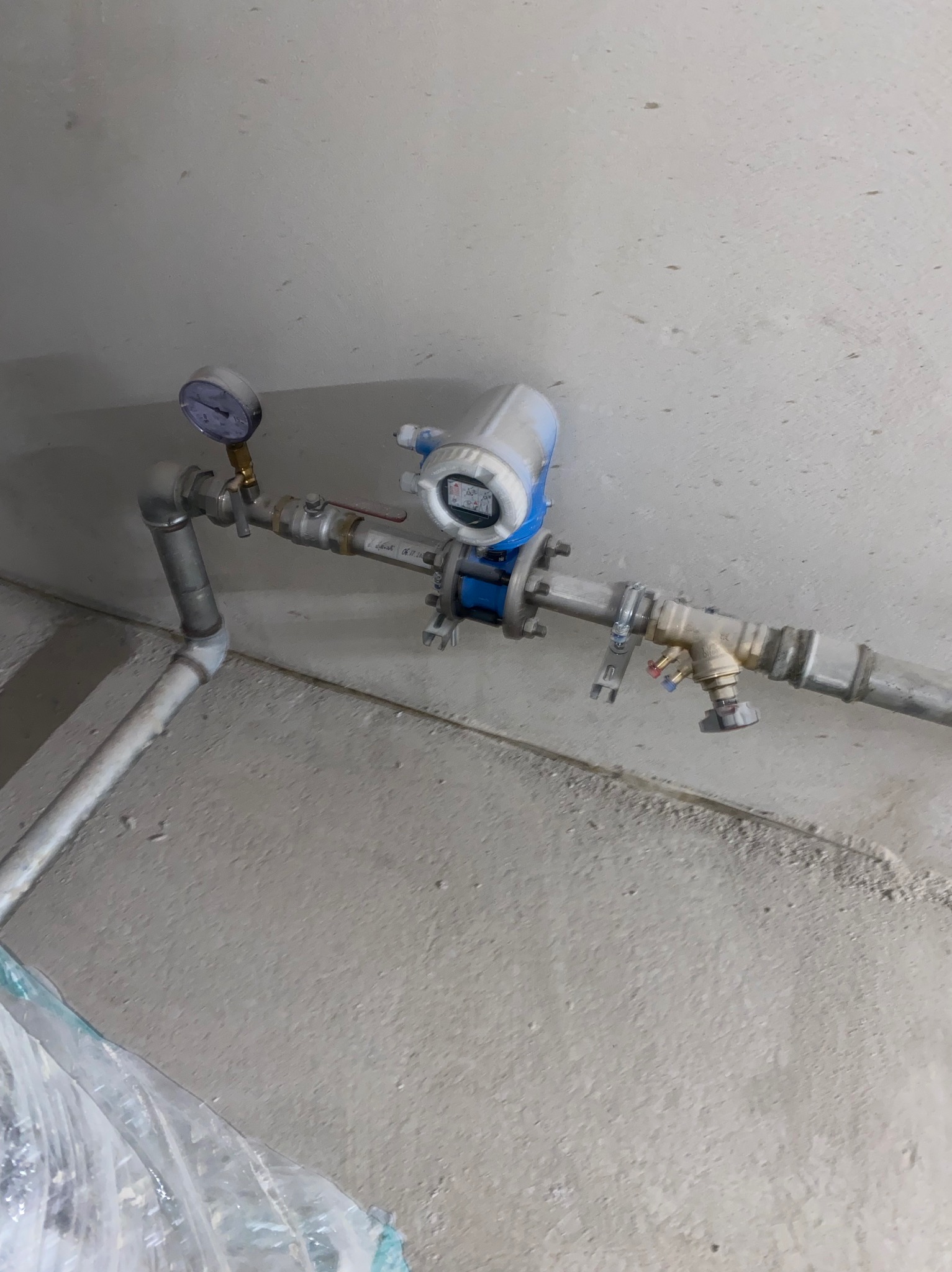 Szara instalacja hydrauliczna z manometrem i zaworami, zamontowana przy ścianie. Widoczne elementy złączek i rur.