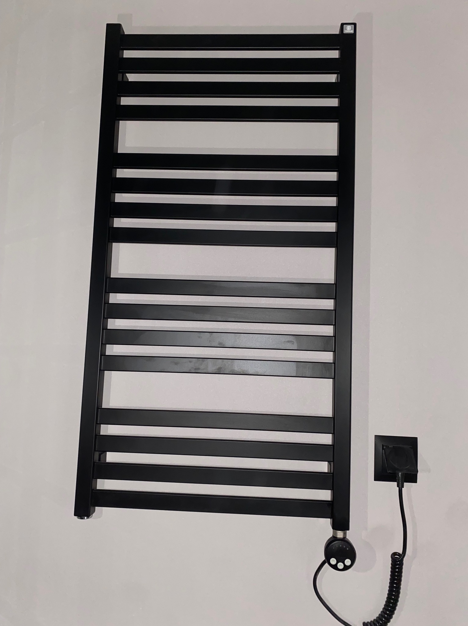Czarny, elektryczny grzejnik łazienkowy drabinkowy, zamontowany na białej ścianie, z widocznym gniazdkiem i spiralnym kablem zasilającym.