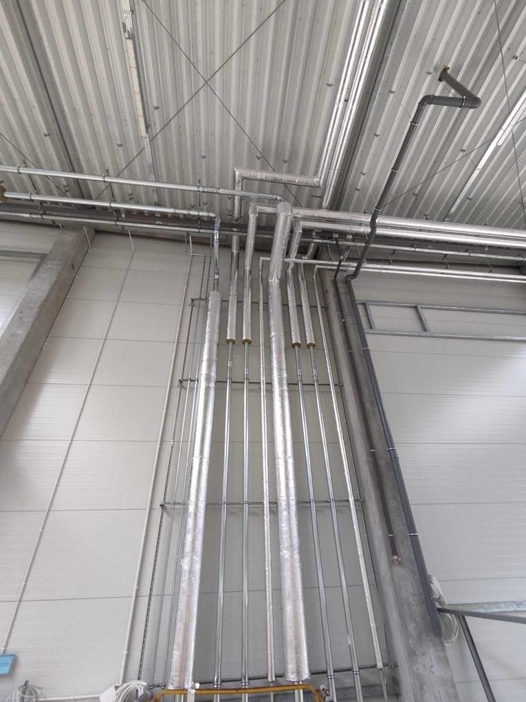 Równoległe rury instalacyjne, niektóre zaizolowane, prowadzone wzdłuż ściany w hali, z widocznymi elementami podwieszenia pod sufitem. Metalowa konstrukcja.