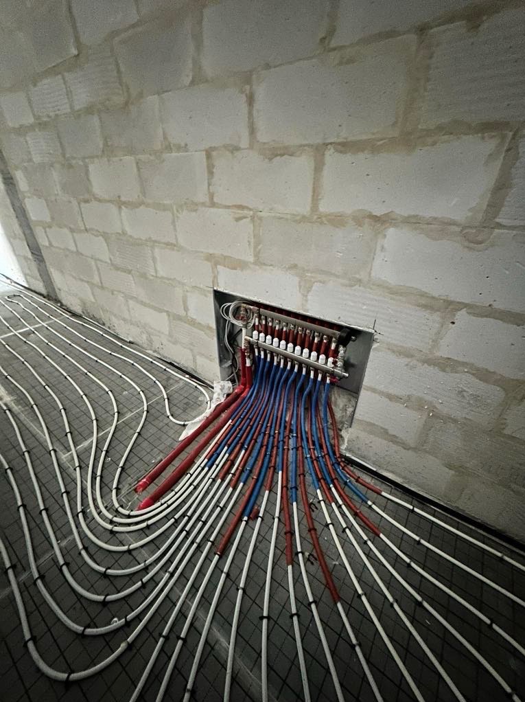 Rozdzielacz ogrzewania podłogowego z rurami rozprowadzonymi na siatce w surowym wnętrzu. Rury białe, niebieskie i czerwone. Ściana z białych bloczków.