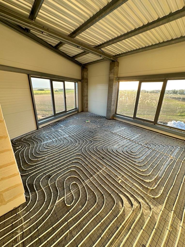 Białe rury ogrzewania podłogowego ułożone na siatce w jasnym pomieszczeniu z oknami i widokiem na pole. Metalowa konstrukcja dachu, surowy stan wykończenia.