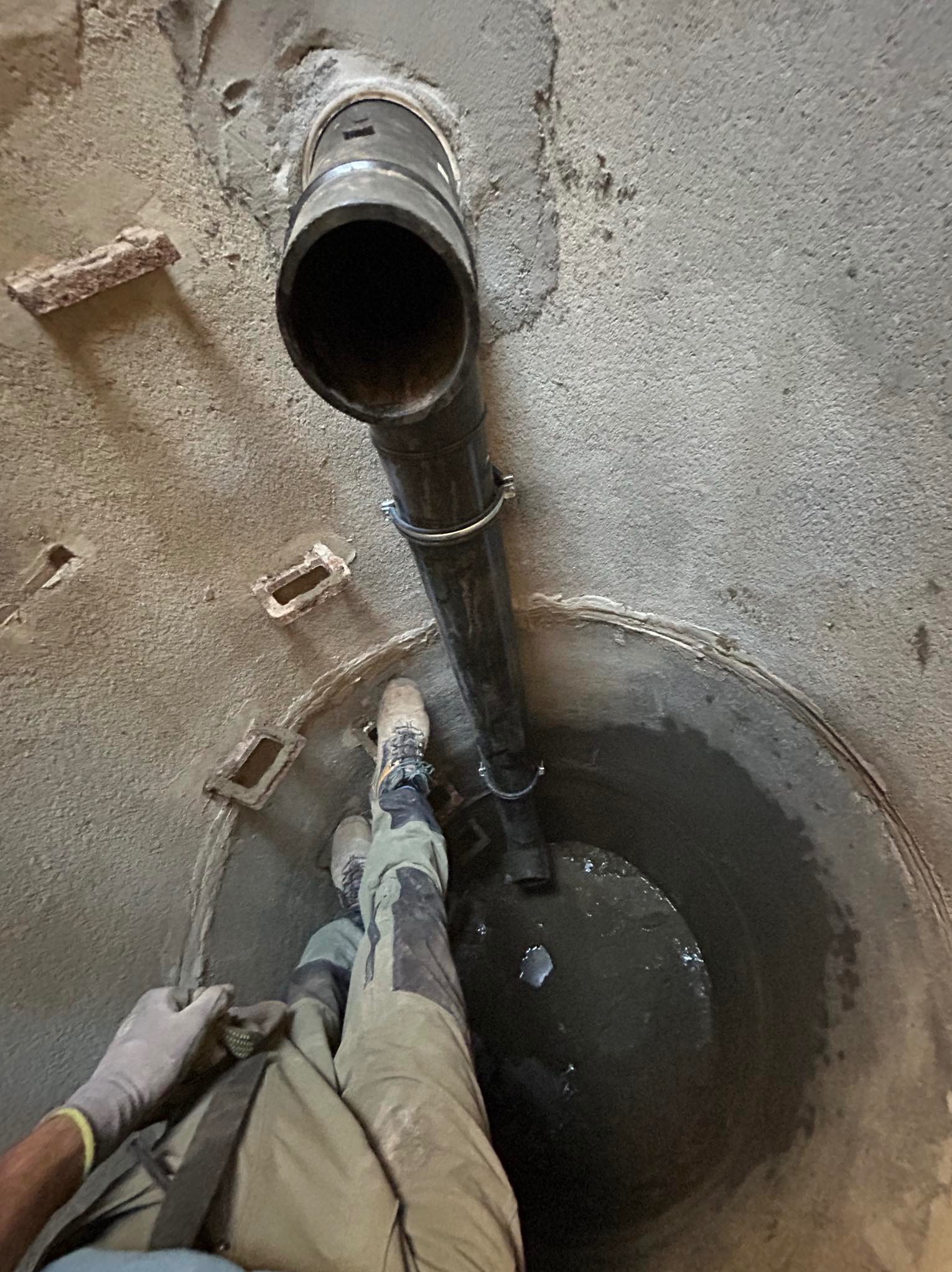 Wnętrze studni kanalizacyjnej z zamontowaną pionową rurą odpływową, zabezpieczoną obejmami. Widoczne dno studni z wodą i fragment sylwetki pracownika w odzieży roboczej.