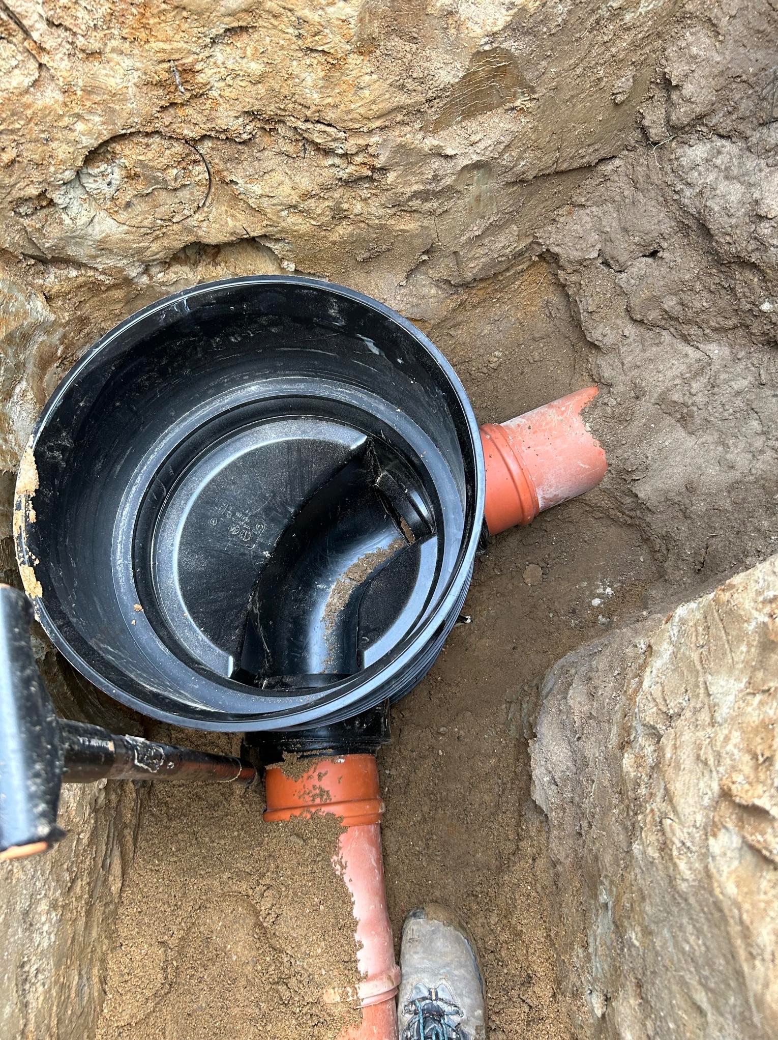 Czarna studzienka kanalizacyjna z rurami w wykopie, widok z góry. Element instalacji kanalizacyjnej w ziemi, pomarańczowe rury. But na dole kadru.