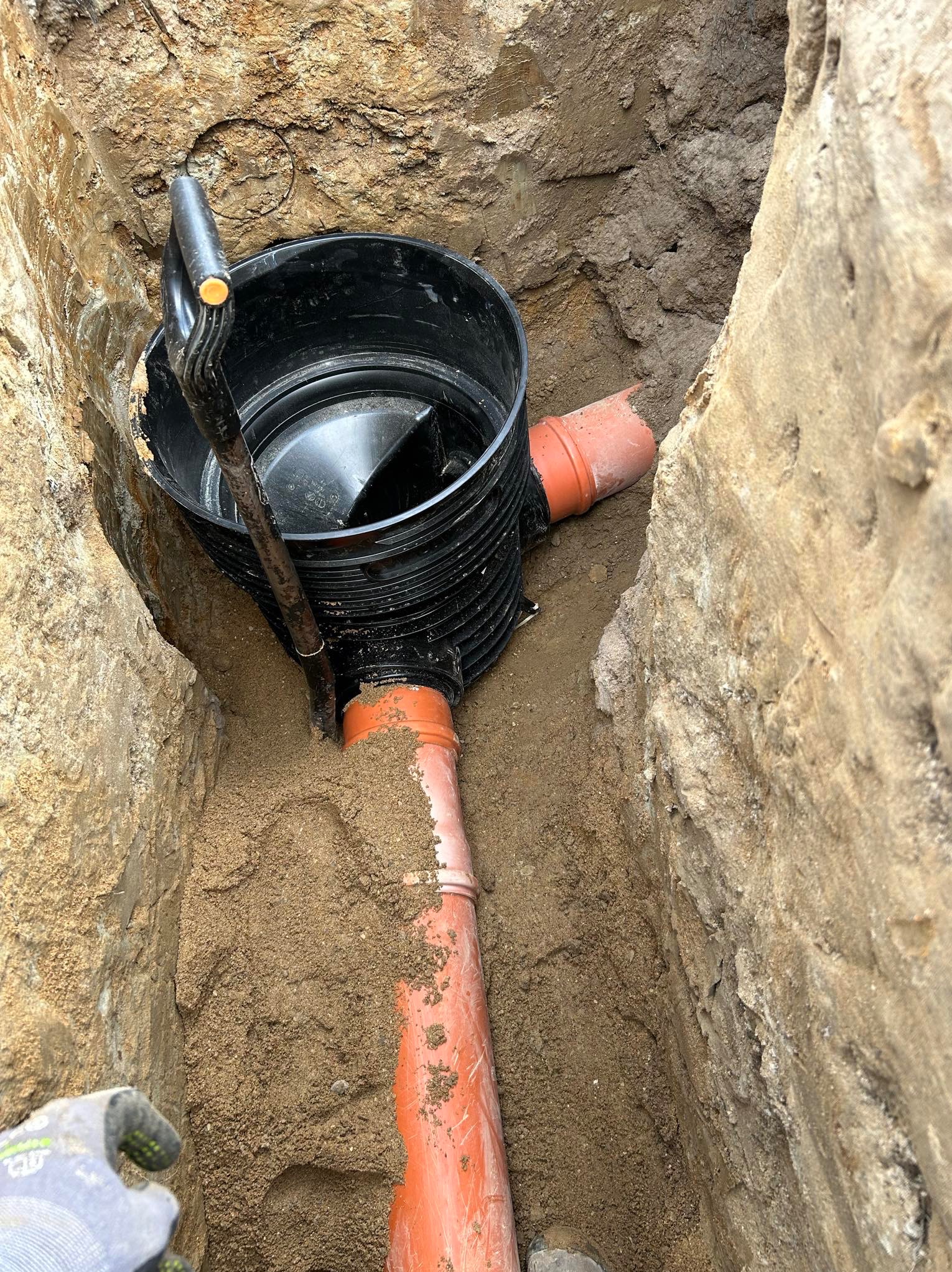 Studzienka kanalizacyjna z rurami w wykopie ziemnym, widok z góry. Łopata oparta o studzienkę. Montaż przyłącza wod-kan w trakcie realizacji.