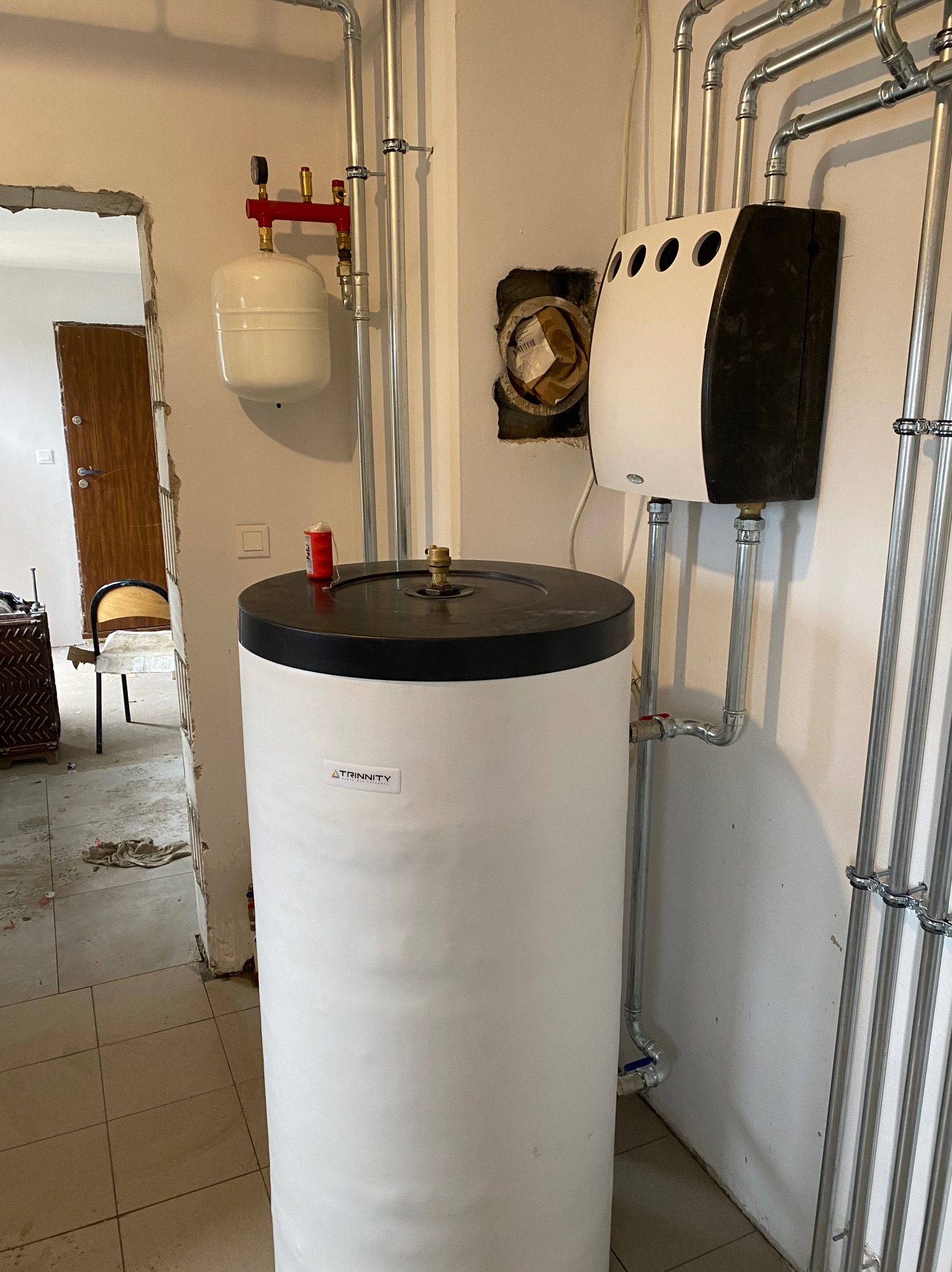 Biały bojler marki Trinity z czarnym panelem, zainstalowany w pomieszczeniu z widocznymi rurami instalacji grzewczej i fragmentem niedokończonego przejścia.