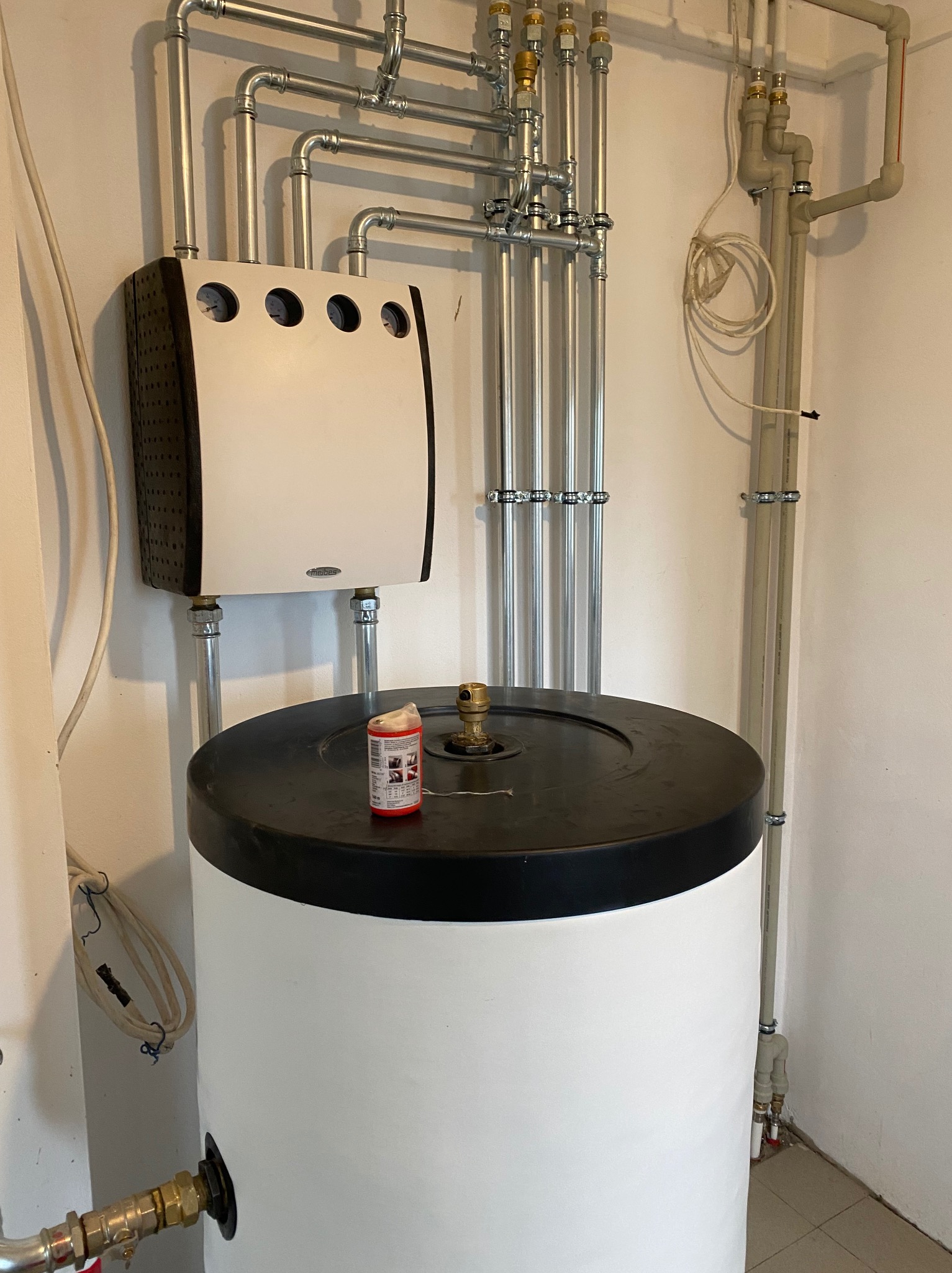 Biały bojler z czarną pokrywą, instalacja z rur w tle, na górze bojlera stoi czerwona puszka. Widoczne połączenia i zawory. Ujęcie z bliska na instalację.