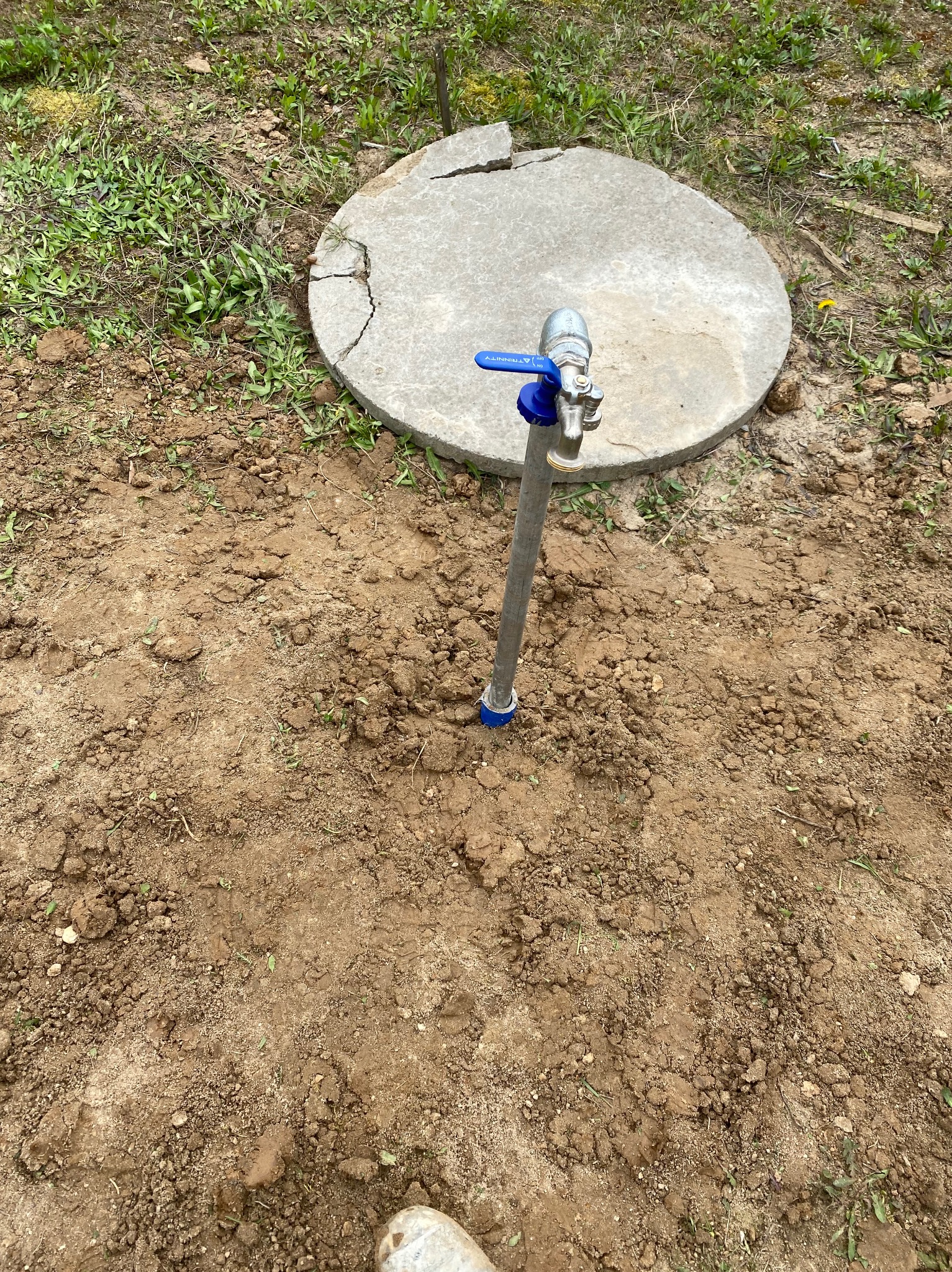 Zewnętrzny kran ogrodowy z niebieskim zaworem, umieszczony na ziemi obok betonowej pokrywy studni. Ziemia wokół świeżo przekopana, widoczne ślady ingerencji.