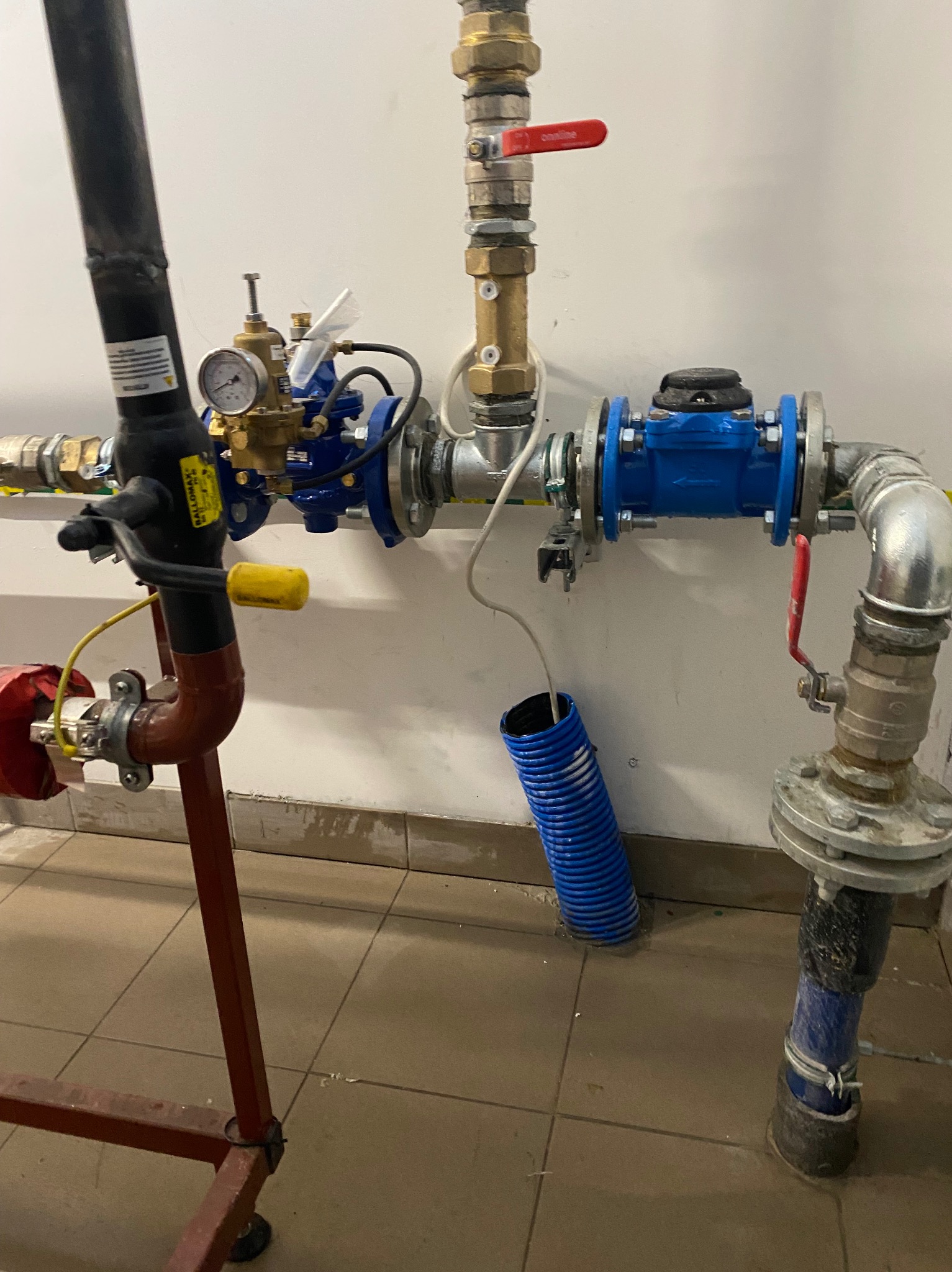 Złożony układ instalacji wodnej z rurami miedzianymi, stalowymi i plastikowymi, zaworami, manometrem, złączkami flanszowymi i elementami mocującymi przy białej ścianie.