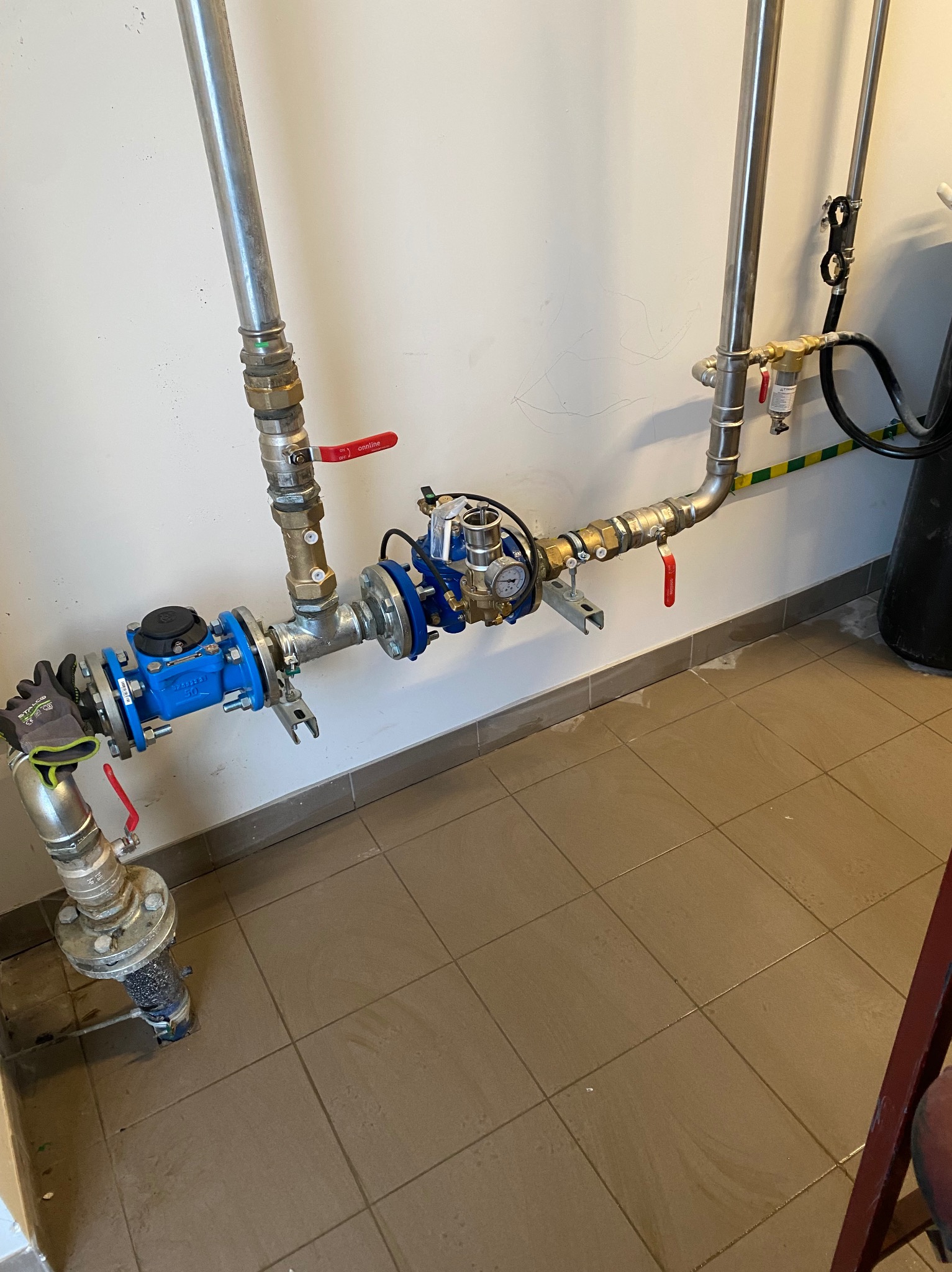 Złożona instalacja wodna z rurami stalowymi, zaworami z czerwonymi uchwytami, niebieskim licznikiem i manometrem, zamontowana przy białej ścianie z brązowymi płytkami podłogowymi.