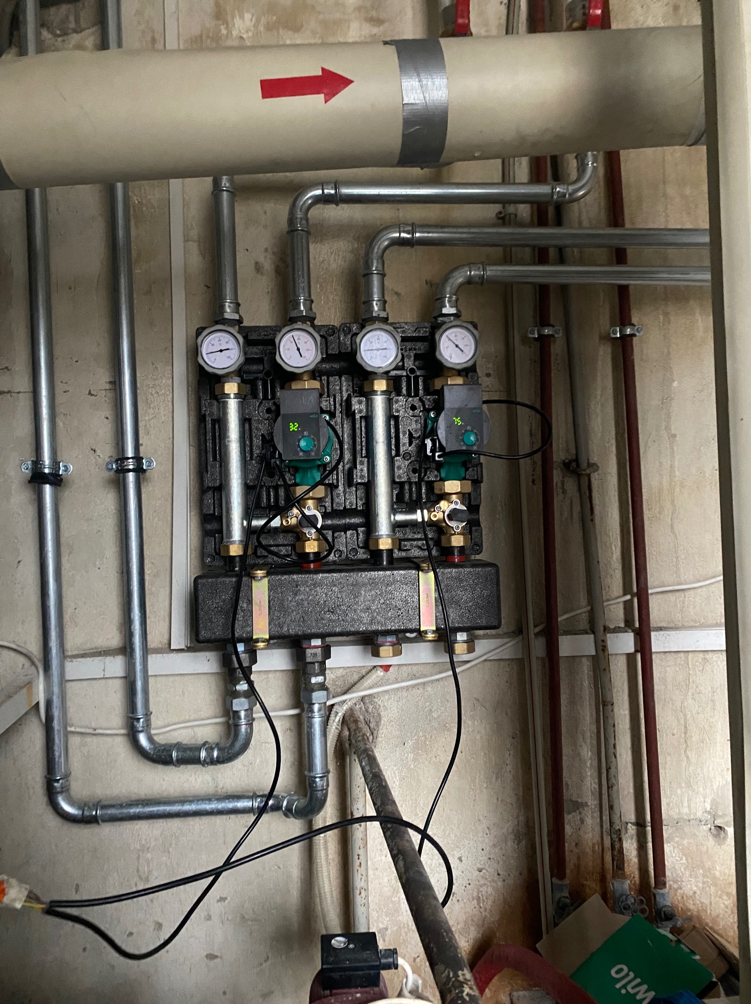 Rozdzielacz ogrzewania podłogowego z czterema obiegami, każdy z manometrem i pompą obiegową, zamontowany na ścianie z rurami doprowadzającymi i odprowadzającymi wodę.