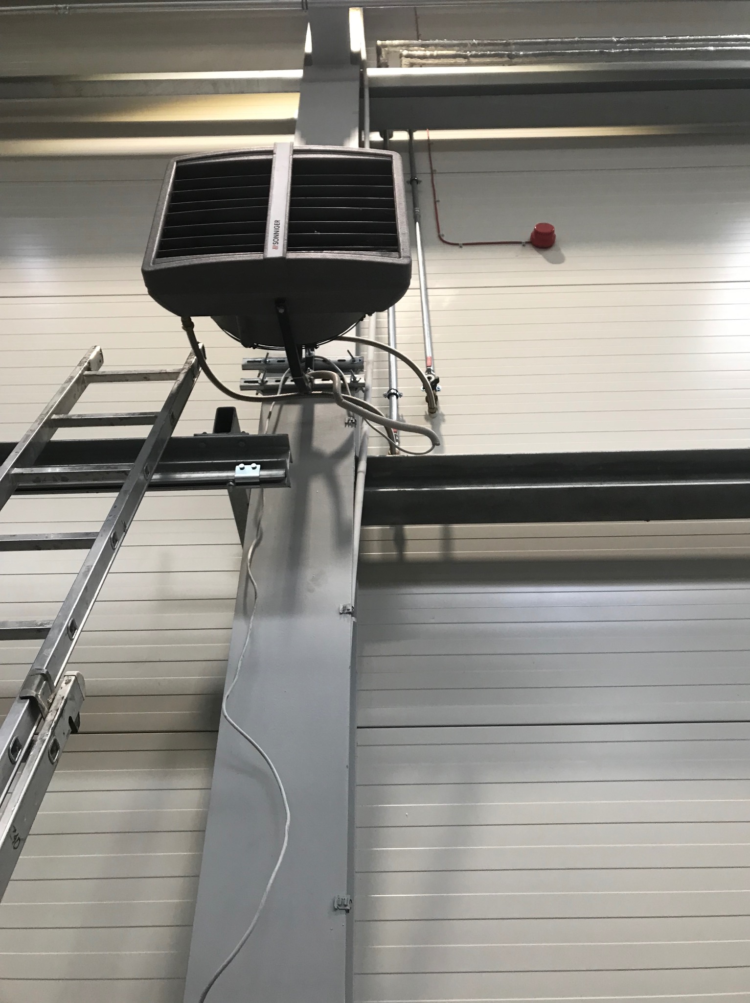 Szary nagrzewnica powietrza Sonniger zamontowana na metalowej kolumnie, drabina oparta o kolumnę, instalacja elektryczna i przeciwpożarowa na ścianie z paneli.