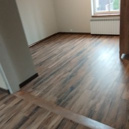 Krystian Zięba KZ HOME Usługi Remontowo Wykończeniowe - Jasne wnętrze z panelami podłogowymi w odcieniach brązu, widok na okno z grzejnikiem. Ściany w neutralnym kolorze, sufit z lampą. Część szafy po prawej.