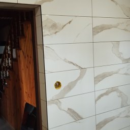 Krystian Zięba KZ HOME Usługi Remontowo Wykończeniowe - Wykończona ściana w łazience z białych płytek imitujących marmur z brązowymi żyłami. Widoczny fragment drewnianych schodów i narzędzia na podłodze.