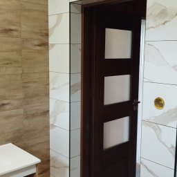 Krystian Zięba KZ HOME Usługi Remontowo Wykończeniowe - Wykończona łazienka z płytkami imitującymi drewno i marmur, widoczna drewniana szafka z białym blatem oraz ciemne drzwi z przeszkleniami.