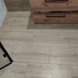 Krystian Zięba KZ HOME Usługi Remontowo Wykończeniowe - Ukończona łazienka z drewnopodobną podłogą, fragment kabiny prysznicowej i szafka pod umywalką z drewnianymi frontami. Widok z góry, neutralne oświetlenie.