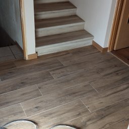 Krystian Zięba KZ HOME Usługi Remontowo Wykończeniowe - Ułożone płytki podłogowe imitujące drewno, prowadzące do schodów z jasnymi stopniami. Widoczny fragment narzędzia pracy w rogu kadru.