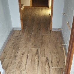 Krystian Zięba KZ HOME Usługi Remontowo Wykończeniowe - Ułożona podłoga z płytek imitujących drewno w korytarzu, widok z progu. Jasne ściany, drewniane futryny. W tle widoczny telewizor i meble.