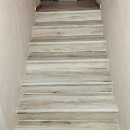 Krystian Zięba KZ HOME Usługi Remontowo Wykończeniowe - Wykończone schody z panelami imitującymi drewno, prowadzące do góry. Ściany w jasnym odcieniu beżu. Widoczne detale wykończenia i faktura paneli.