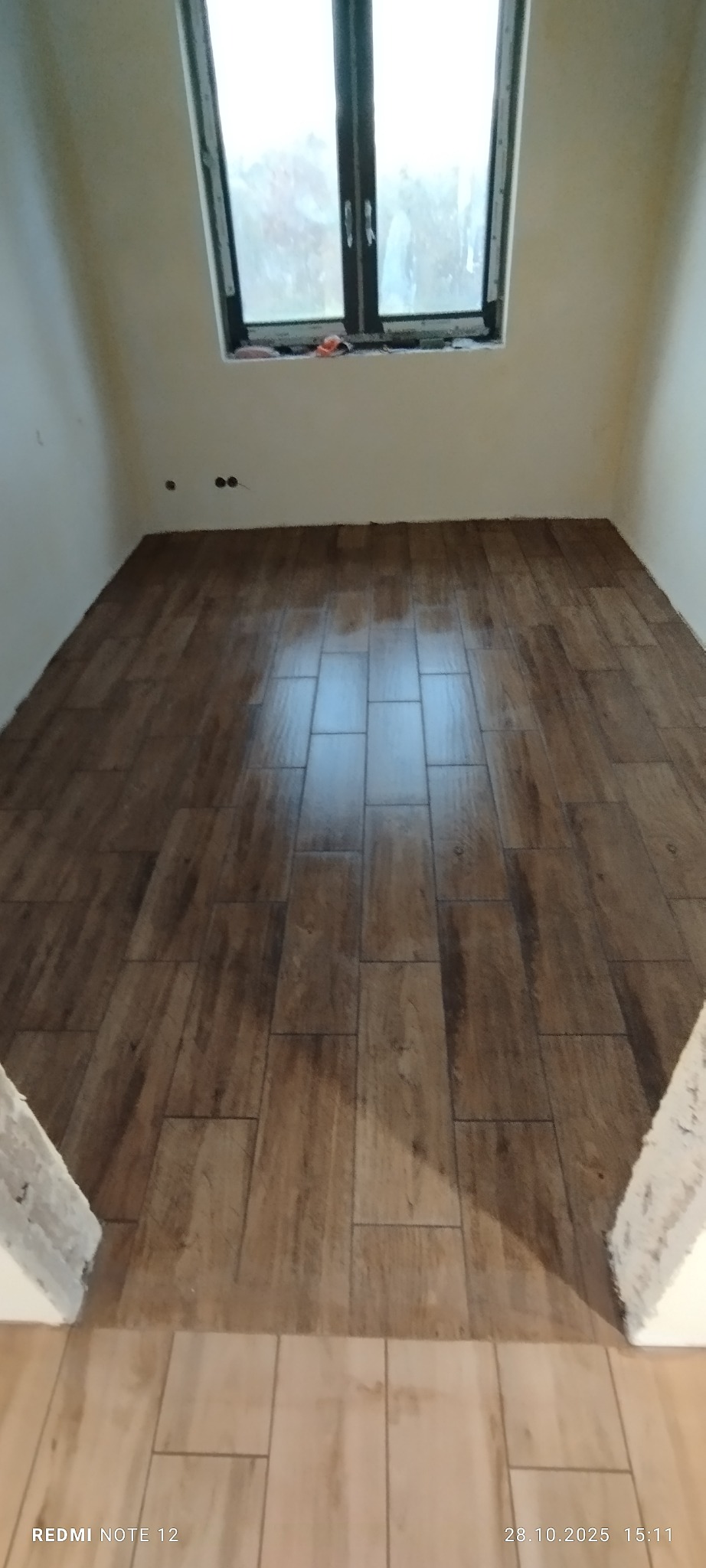 Ukończona podłoga z płytek imitujących drewno w małym pomieszczeniu z oknem. Widoczne fugi i fragment progu z jasnymi płytkami. Ściany w kolorze złamanej bieli.