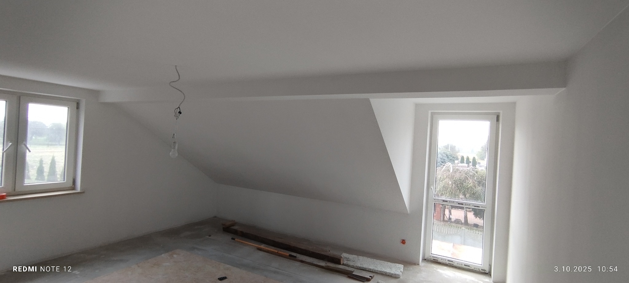 Białe, świeżo pomalowane poddasze z oknem i widocznym okablowaniem. Na podłodze leżą deski. Minimalistyczne wykończenie wnętrza w jasnych barwach.