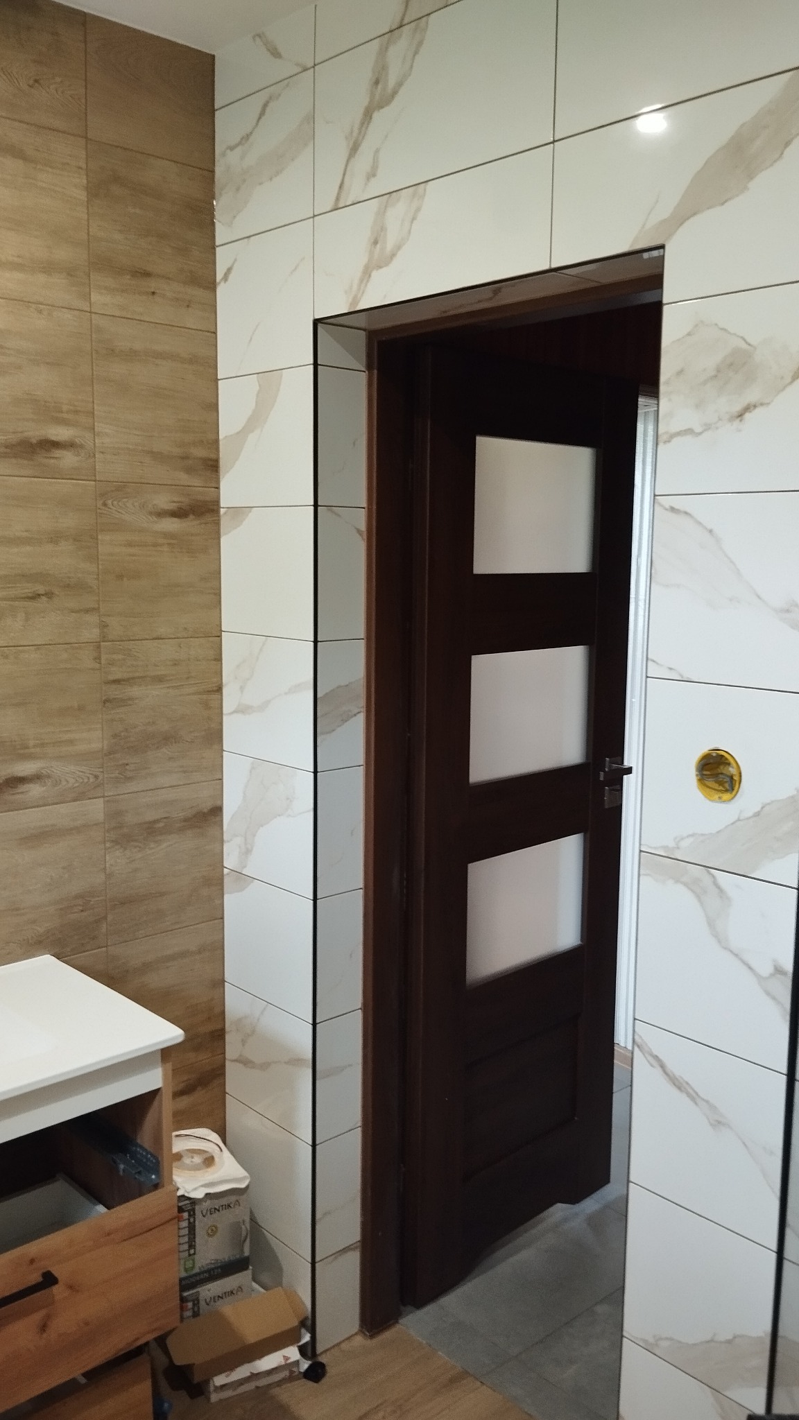 Wykończona łazienka z płytkami imitującymi drewno i marmur, widoczna drewniana szafka z białym blatem oraz ciemne drzwi z przeszkleniami.