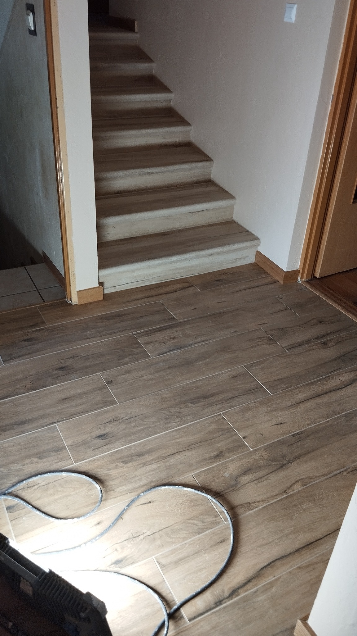Ułożone płytki podłogowe imitujące drewno, prowadzące do schodów z jasnymi stopniami. Widoczny fragment narzędzia pracy w rogu kadru.