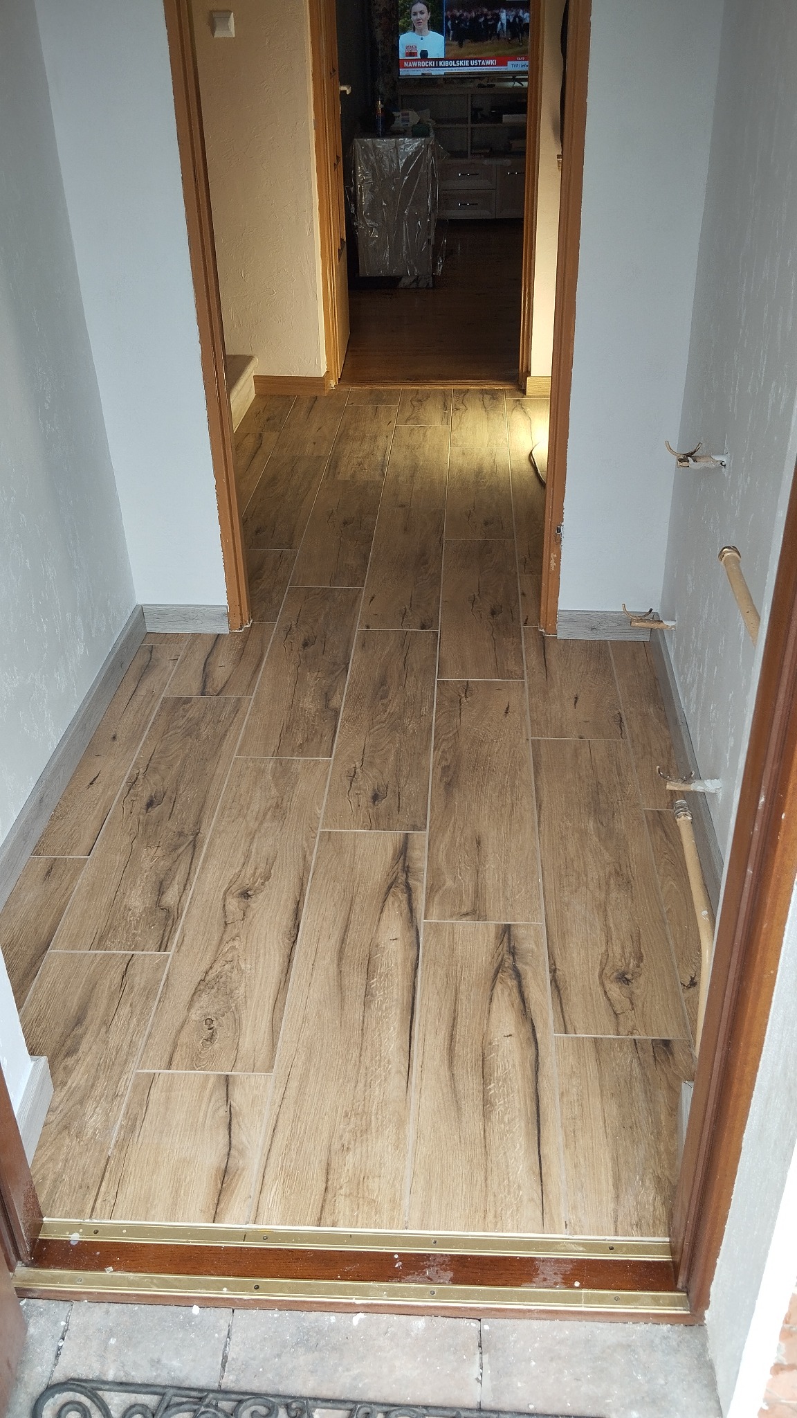 Ułożona podłoga z płytek imitujących drewno w korytarzu, widok z progu. Jasne ściany, drewniane futryny. W tle widoczny telewizor i meble.